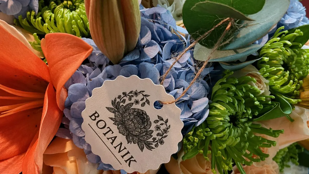 Close de um buquê com lírio laranja, hortênsia azul e crisântemo verde, com a etiqueta da Botanik em primeiro plano.