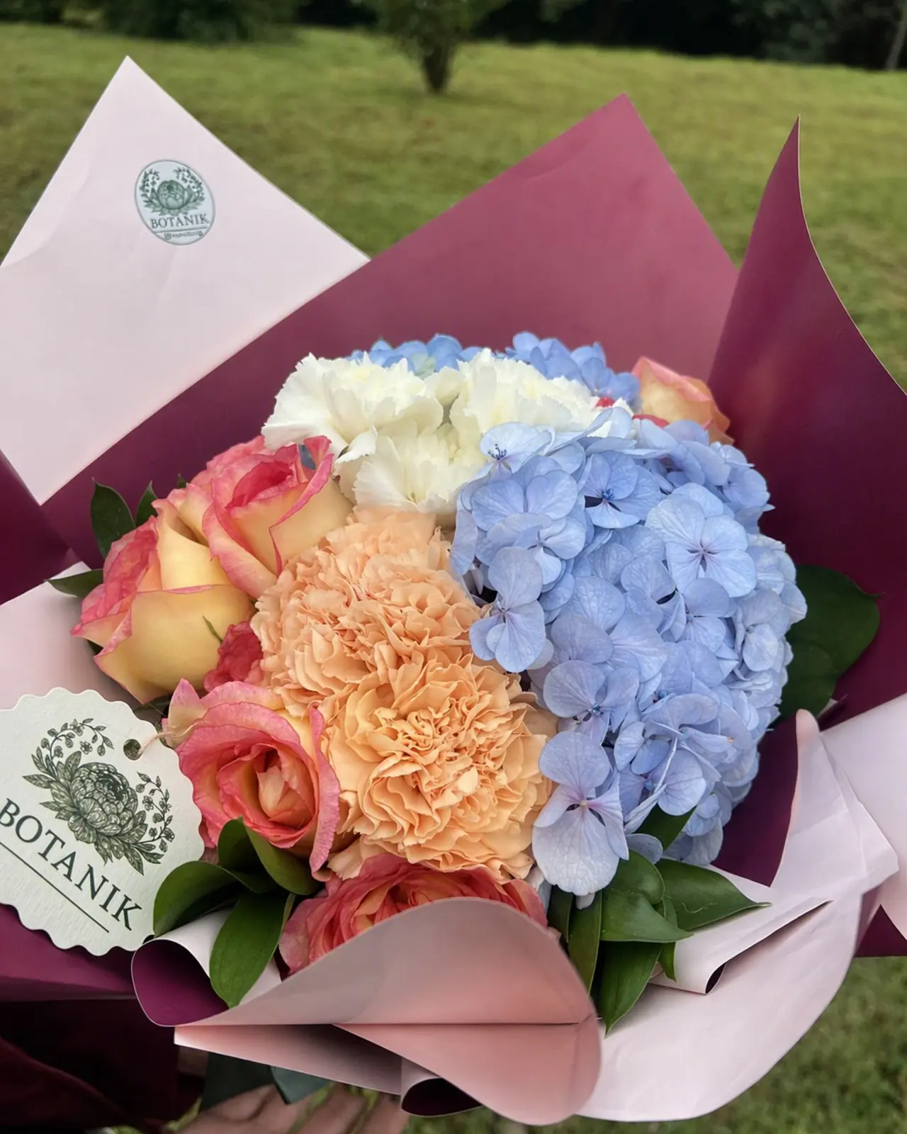 Buquê de hortênsias azuis, rosas pêssego e cravos brancos em papel bordô e rosa.