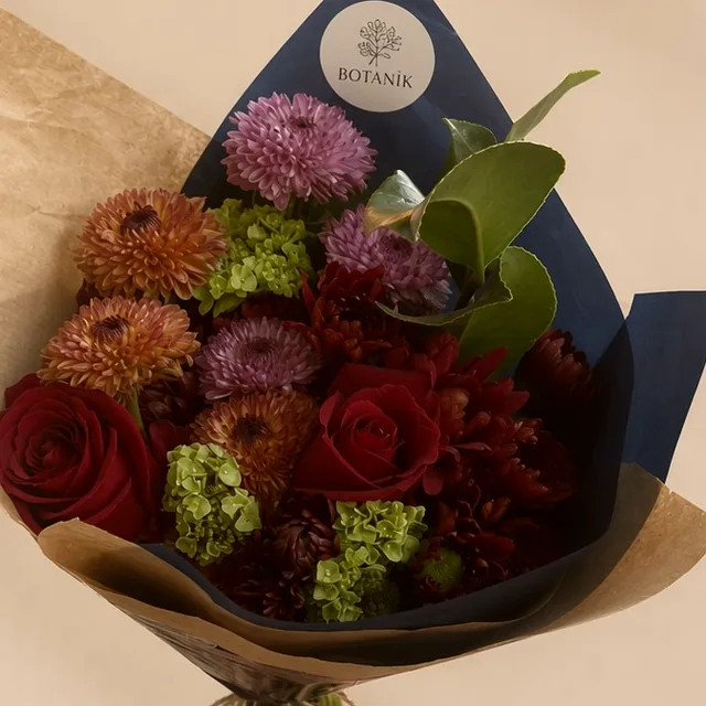 Buquê de rosas bordô, crisântemos e hortênsia verde, embrulhado em papel azul-marinho com o selo da Botanik.