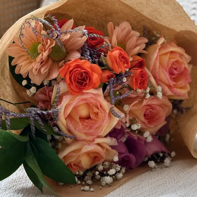 Buquê em tons de pêssego e laranja, com rosas e gipsofila, sobre manta clara.