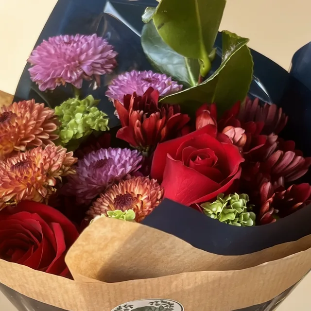 Buquê de rosas vermelhas e crisântemos lilás em papel azul-marinho e kraft.