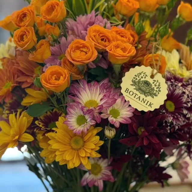 Arranjo alto em tons de laranja e amarelo, com mini-rosas e margaridas, em vaso de vidro.
