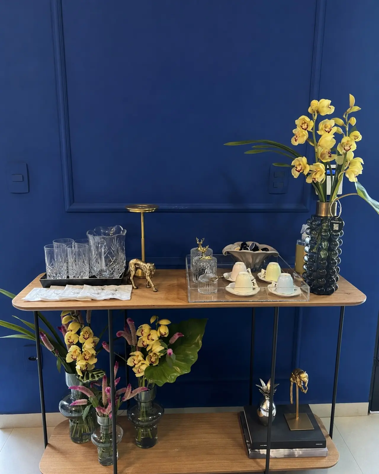 Aparador de madeira com arranjos florais nas duas prateleiras, copos e objetos de decoração, contra parede azul-marinho.
