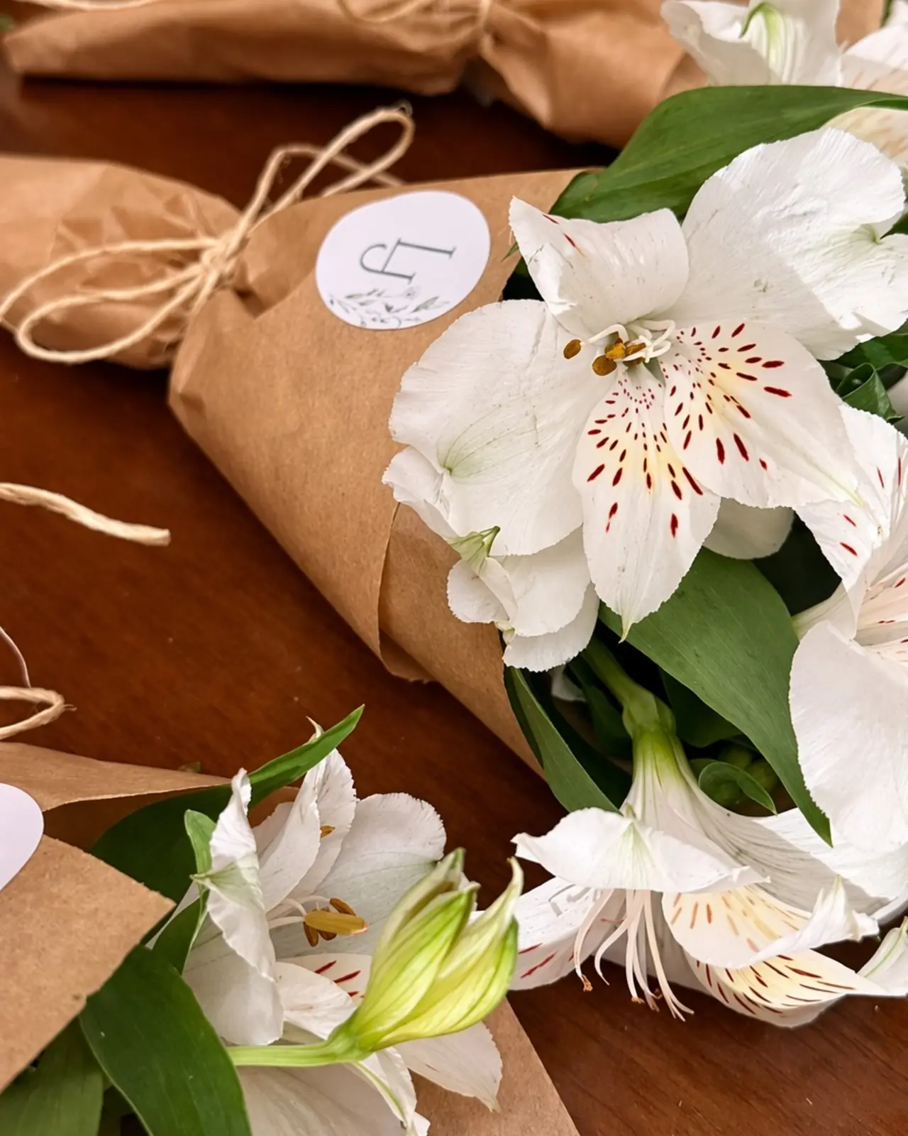 Close de buquês de alstroeméria branca em papel kraft sobre mesa de madeira, com laço de ráfia e a etiqueta da festa.