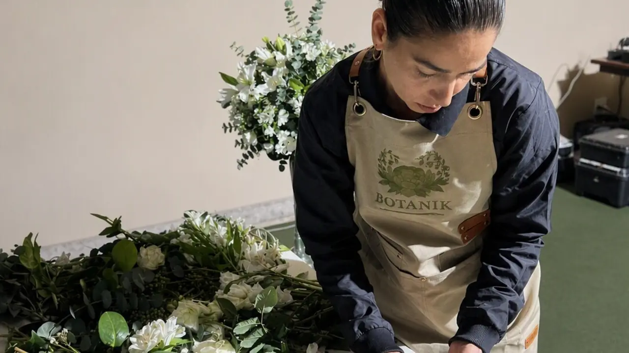 Florista de avental Botanik montando um arranjo sobre a bancada, flores e folhagens em preparo.