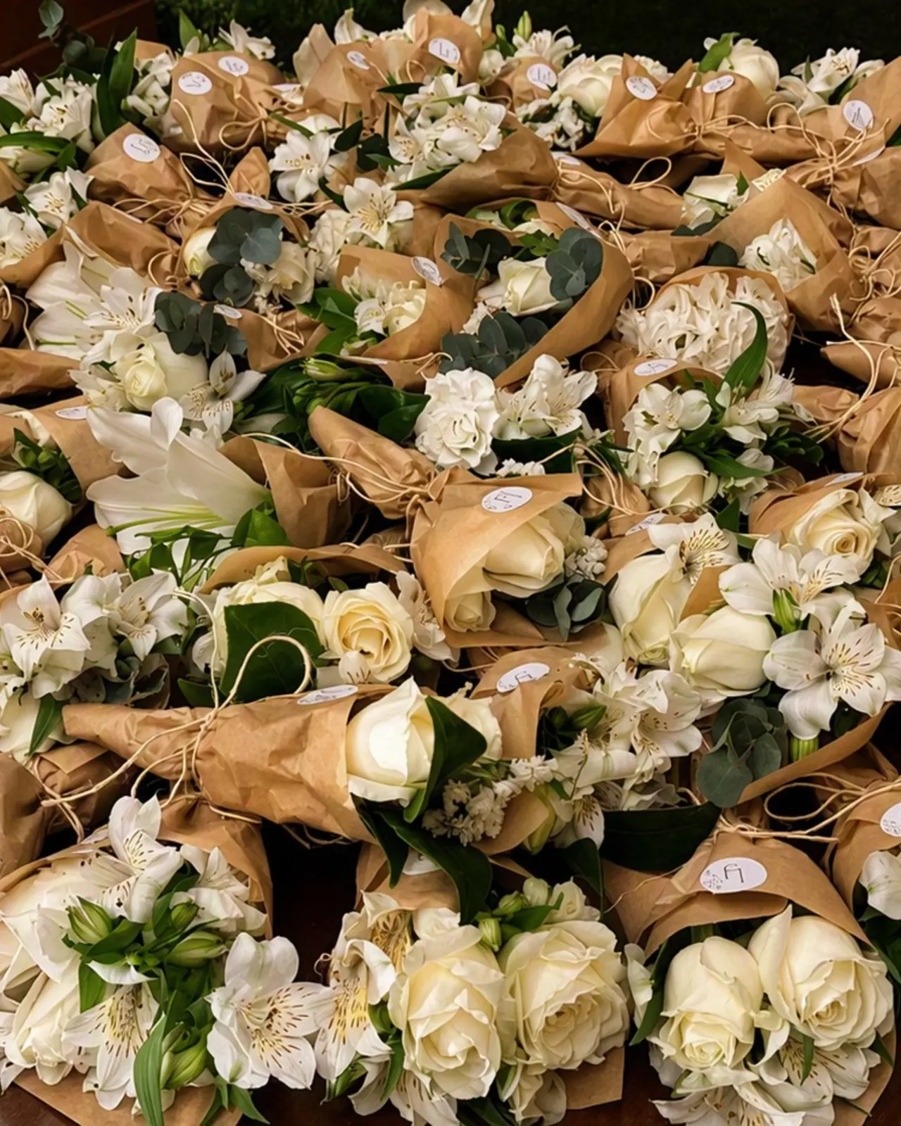 Mesa redonda coberta por dezenas de buquês brancos em papel kraft, cada um com laço de ráfia e etiqueta.
