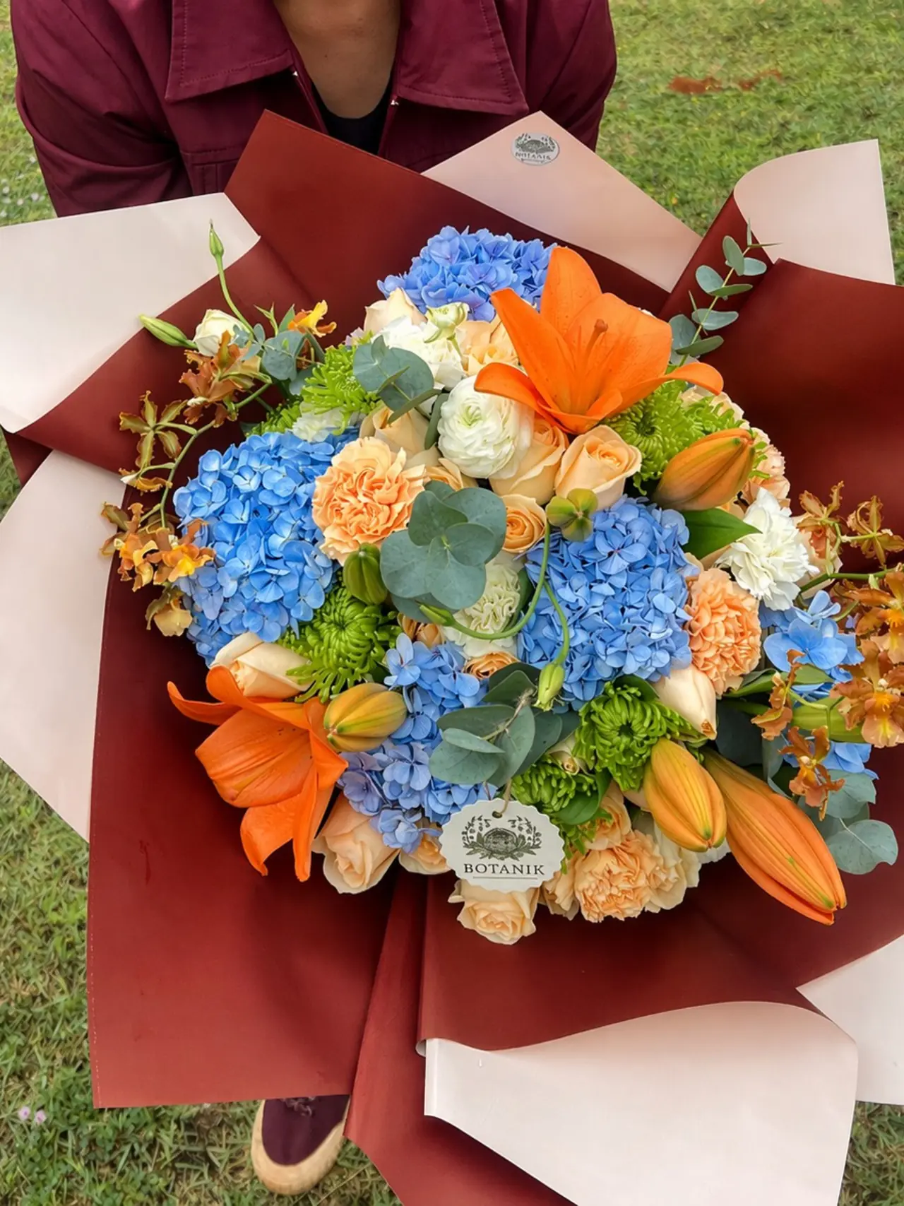 Florista da Botanik segurando um buquê de hortênsias azuis, lírios laranja e rosas em papel bordô, em campo aberto.
